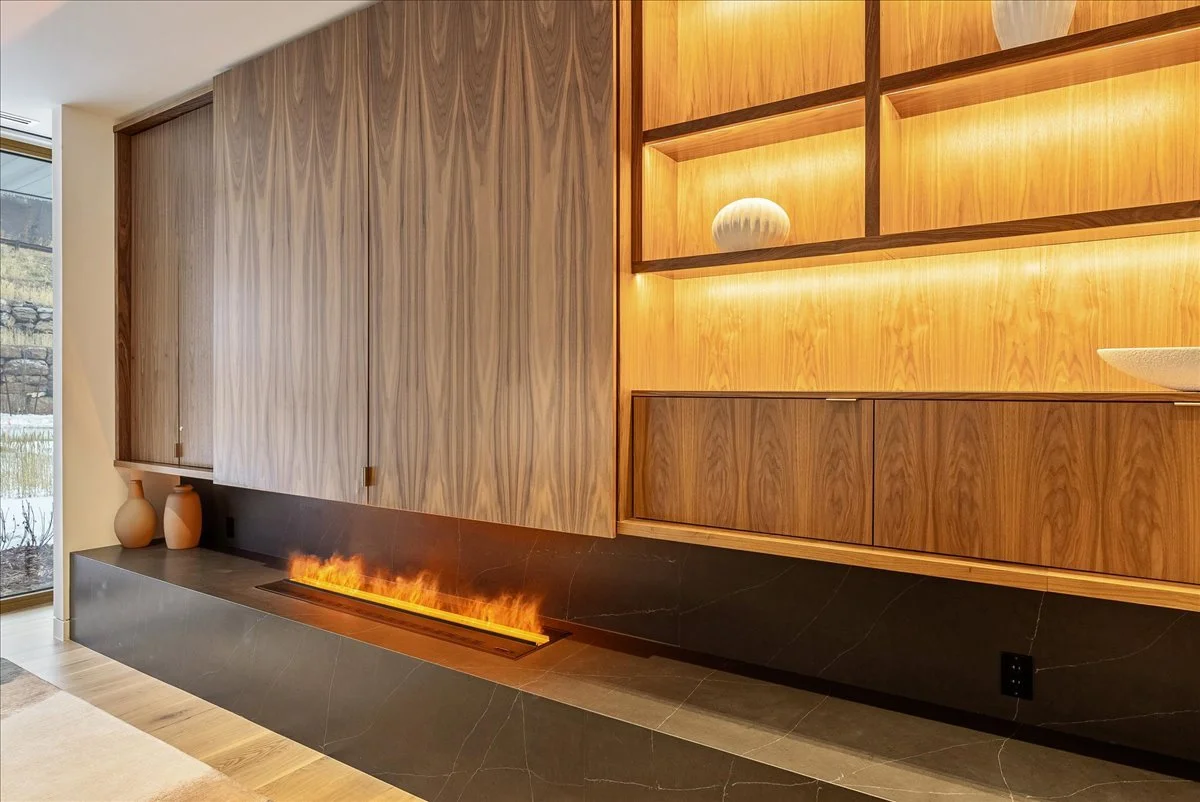 Modern living room with wooden cabinets, a built-in fireplace with flames, decorative vases, and a large window showing an outdoor scene.
