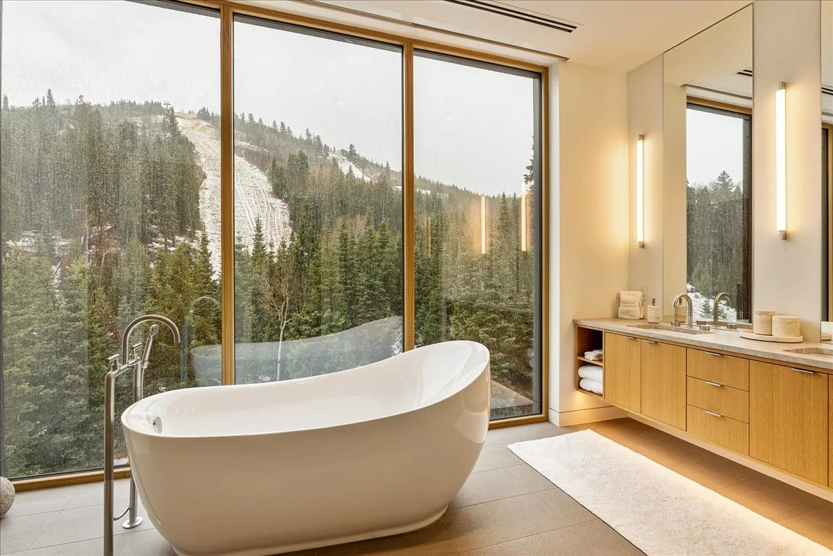 Modern bathroom with a freestanding bathtub near large floor-to-ceiling windows showing a forested mountain landscape.
