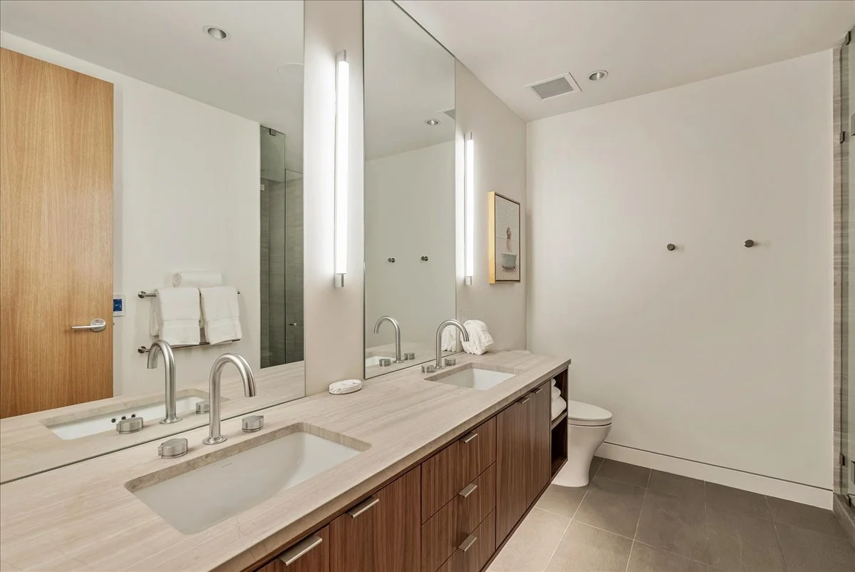 Modern bathroom with double sinks, large mirror, towel rack, toilet, and shower area in the background.