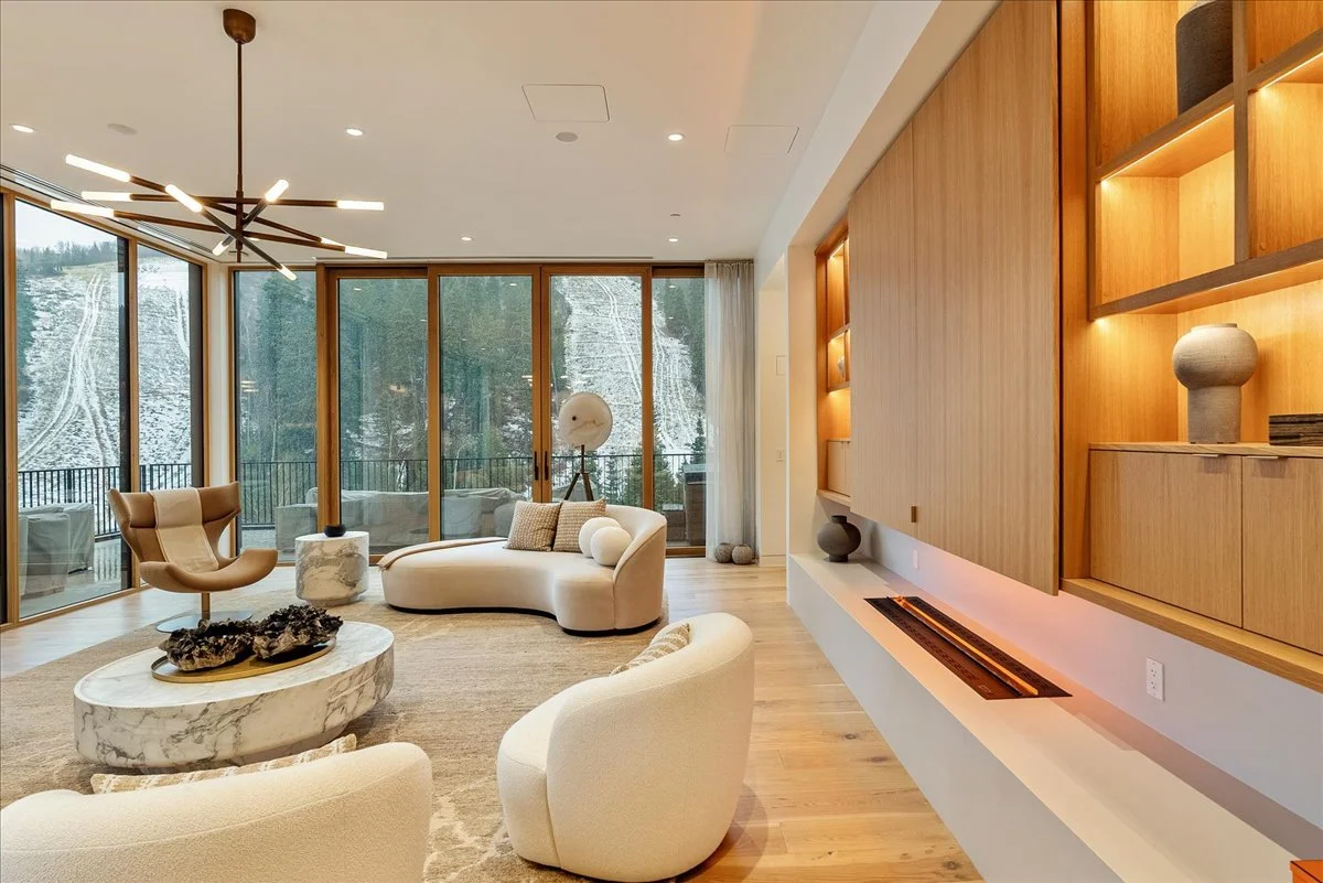 Modern living room with large windows showing snowy mountain landscape, featuring a beige curved sofa, armchairs, marble coffee table, wooden shelving, and contemporary light fixture.