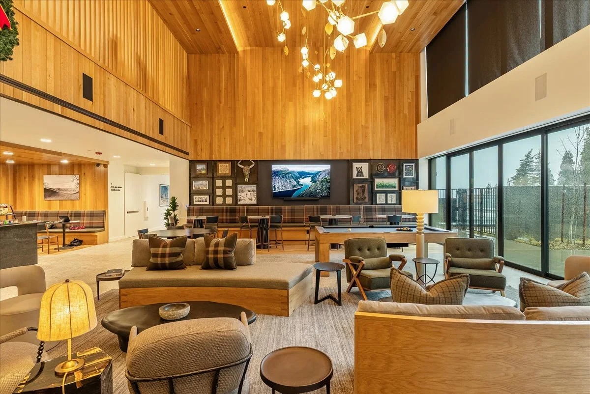 The image shows a spacious, modern living area with wooden accents and large windows. The room is furnished with multiple chairs, sofas, coffee tables, and a TV mounted on a wall. There is warm lighting and a decorative chandelier hanging from the high ceiling.