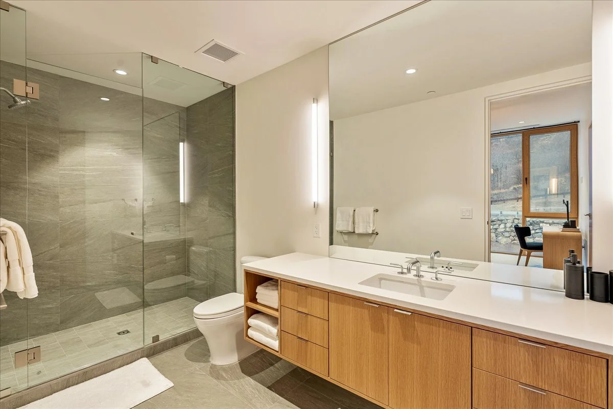 Modern bathroom with a glass-enclosed shower, a white toilet, and a wooden vanity with a white countertop and dual sinks. There are towels on the wall and shelves, and a large mirror. A window with a view of rocks and trees is visible in the backgrou