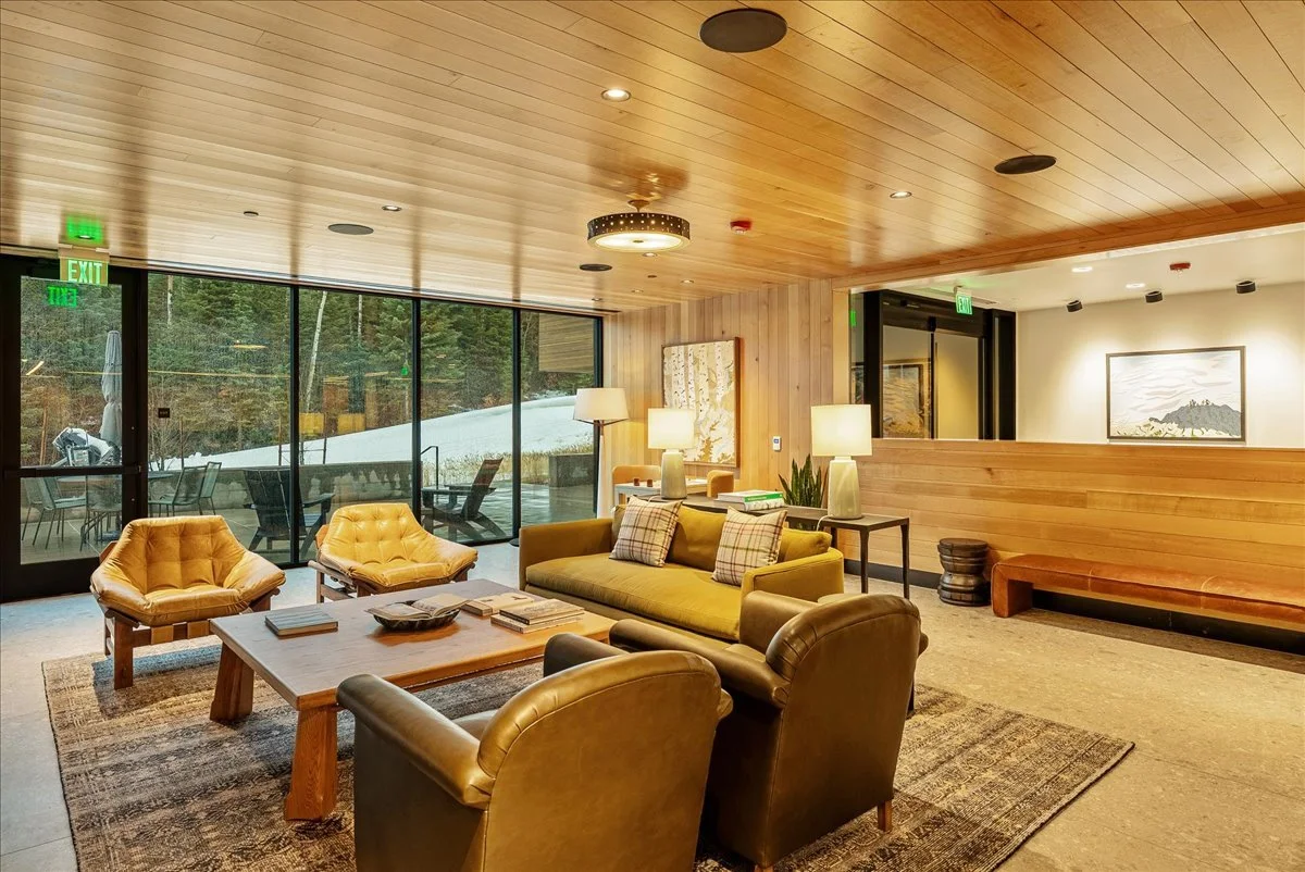 A cozy hotel lobby with a wooden ceiling and walls, large windows showing snow outside, a yellow sofa with plaid pillows, two matching yellow armchairs, a wooden coffee table, side tables with lamps, and a wooden bench along the wall.