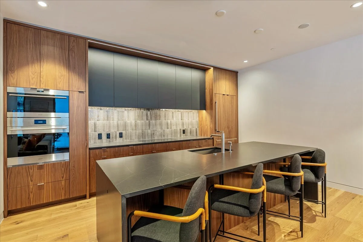 Modern kitchen with wooden cabinetry, a black countertop island with a sink, four gray chairs with wooden armrests, and stainless steel appliances.