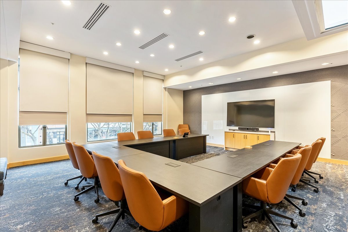Empty conference room with a U-shaped table arrangement, brown swivel chairs, large windows with blinds, and a big flat-screen TV on the wall.