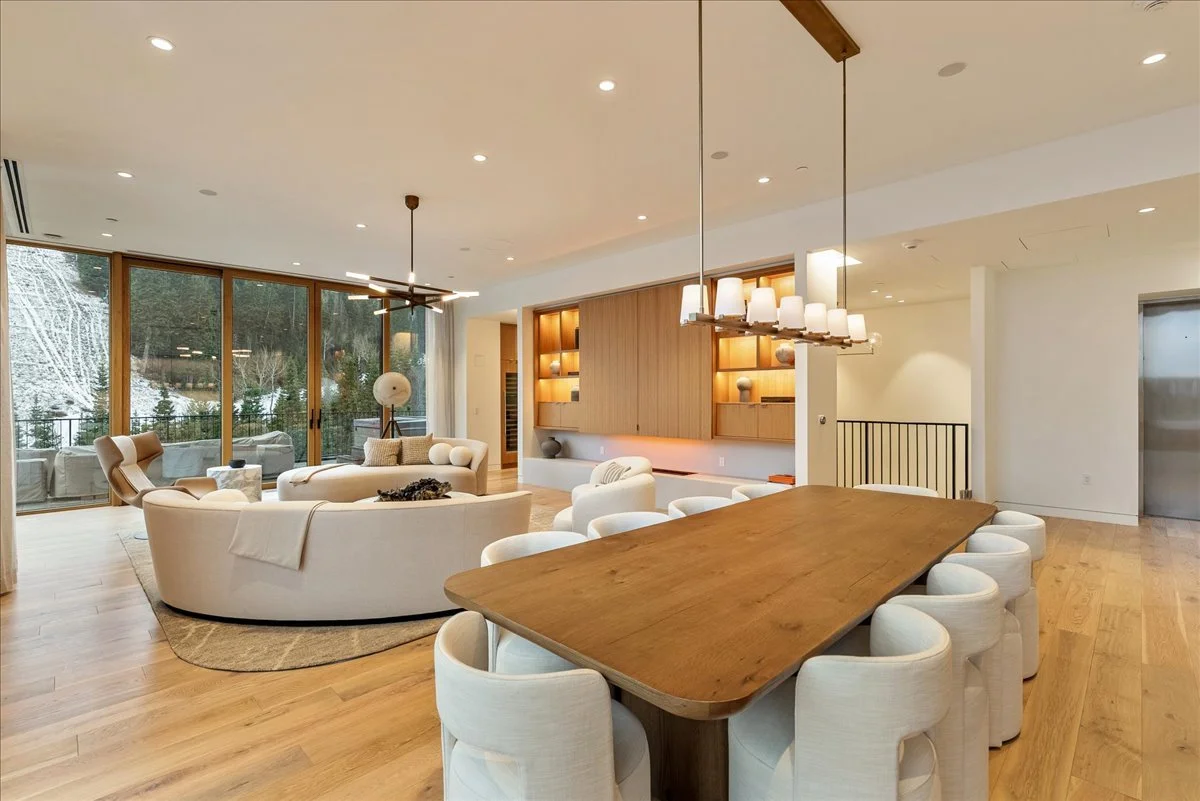 Spacious, modern living and dining area with large windows showing a snowy outdoor landscape, white sofas, wooden dining table with cream chairs, and warm wood accents.