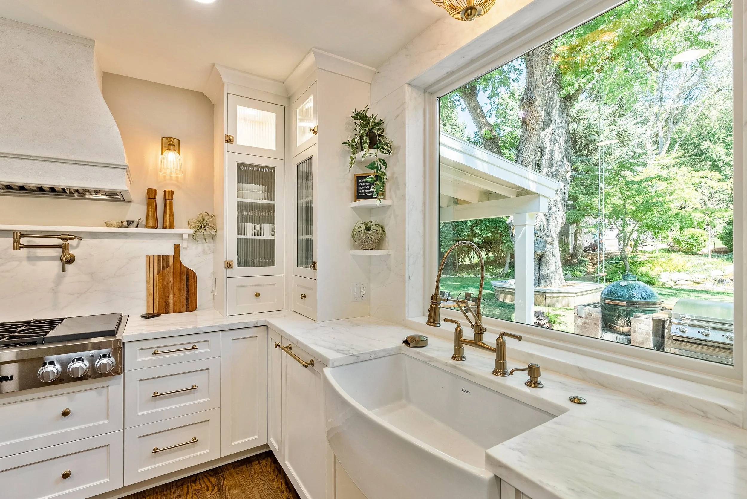 White kitchen with marble countertops and a large window showing a view of green trees outside, a gold faucet, and some decorative items on a shelf.