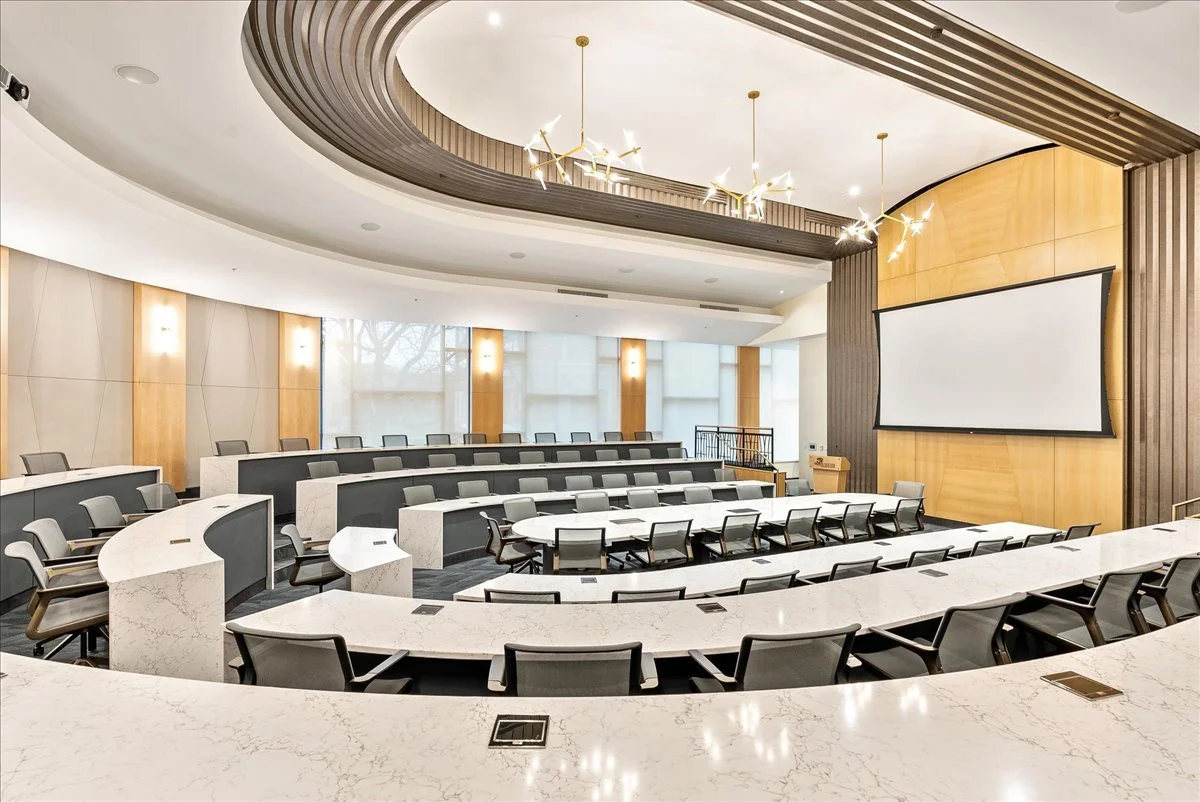 An empty modern conference room with curved tiered seating, a large projection screen, and contemporary lighting fixtures.