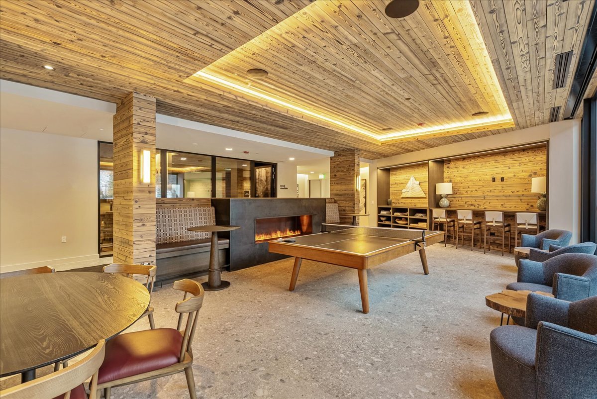 A spacious, modern lounge area with wooden ceiling and walls, featuring a fireplace, ping pong table, seating chairs, a bar counter, and warm lighting.