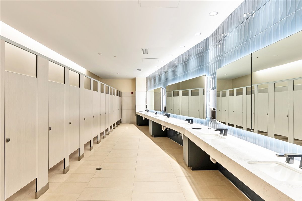 A clean, modern public restroom with white stalls on the left and a long white countertop with multiple sinks and large mirrors on the right.