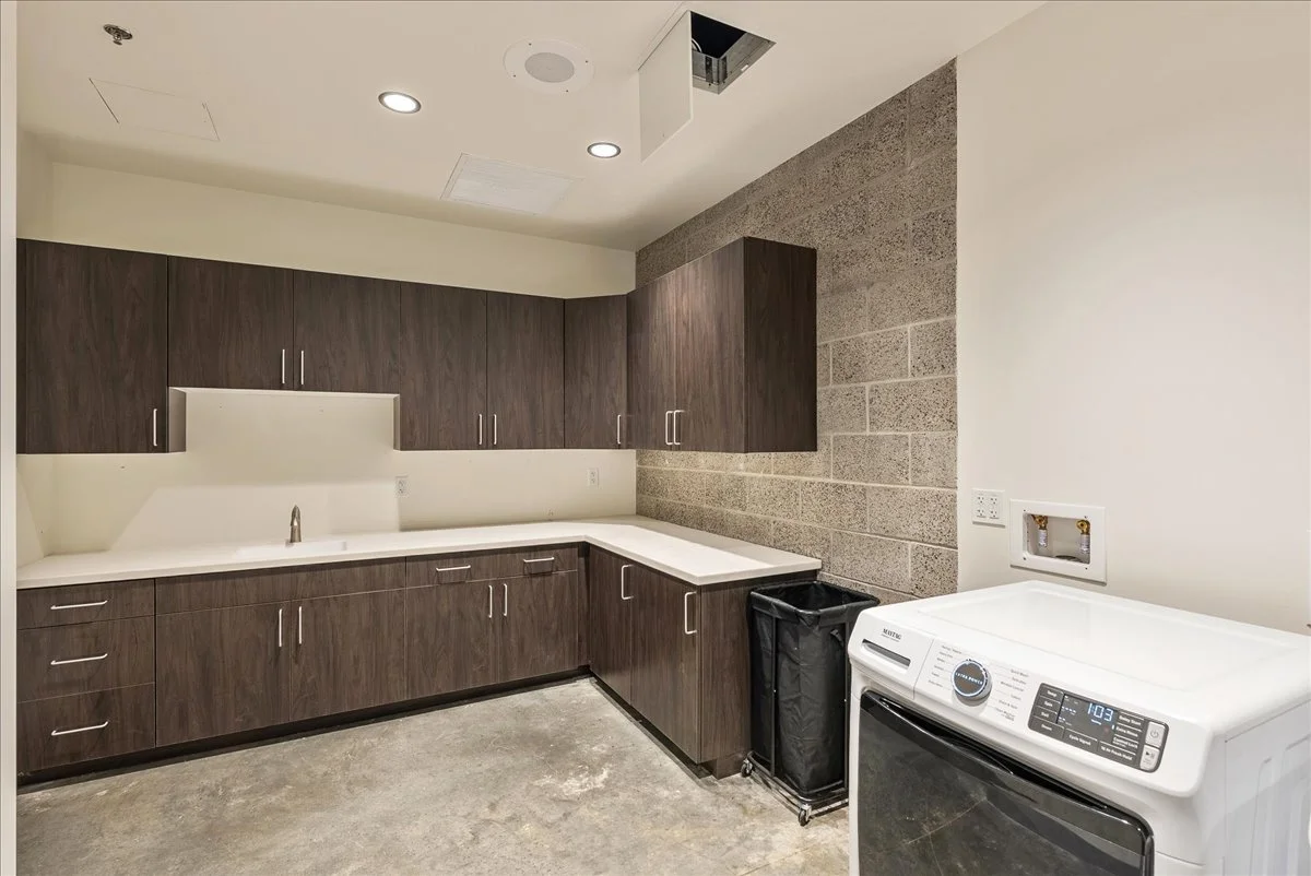 Empty laundry room with brown cabinets, a white countertop, and a washing machine. The room has concrete flooring and a stone accent wall along with unfinished ceiling with lighting.