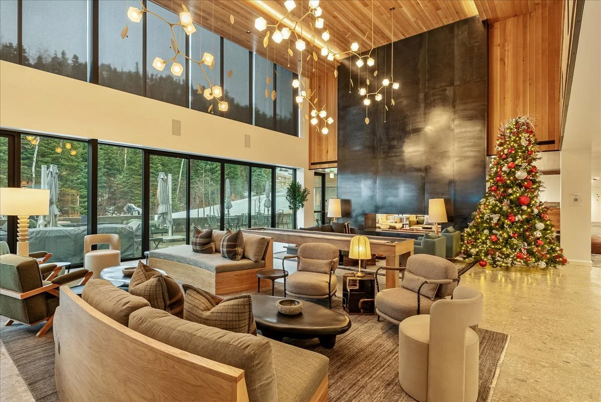 A spacious, modern hotel lobby decorated for Christmas with a Christmas tree adorned with red and silver ornaments and twinkling lights, in front of a large dark wall. The lobby features comfortable seating with various chairs, sofas, and tables, ill