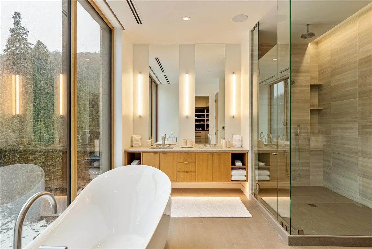 Modern bathroom with a freestanding bathtub, large window, double vanity with mirrors and lighting, and a walk-in glass shower with beige tile walls.