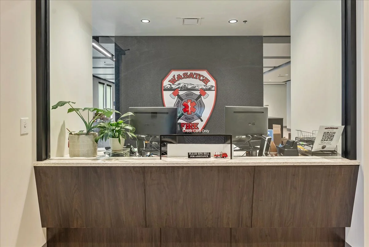 Fire department front desk with two computers, a sign that reads "Please Ring Bell For Assistance," and the fire department logo on the wall behind, with some potted plants on the counter.