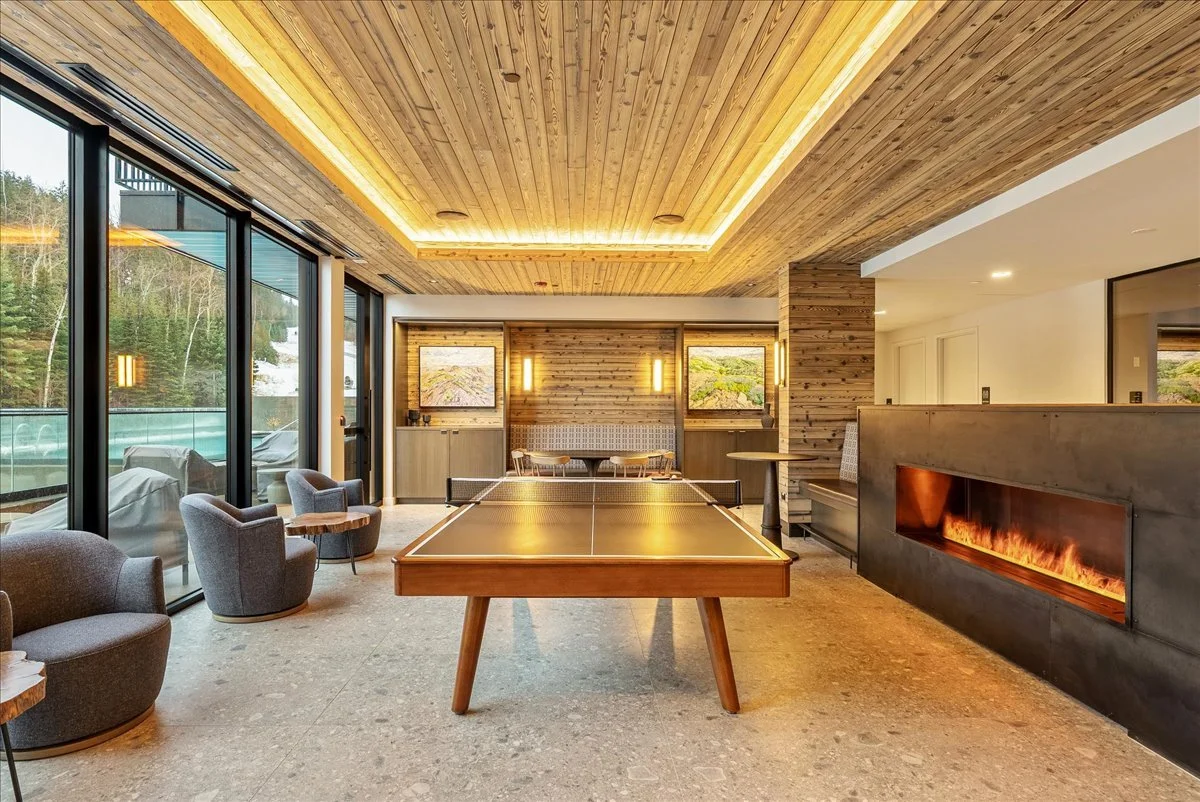 Interior of a modern, cozy lounge with a ping pong table, gray armchairs, a fireplace, wooden ceiling and walls, large glass windows, and outdoor scenery.