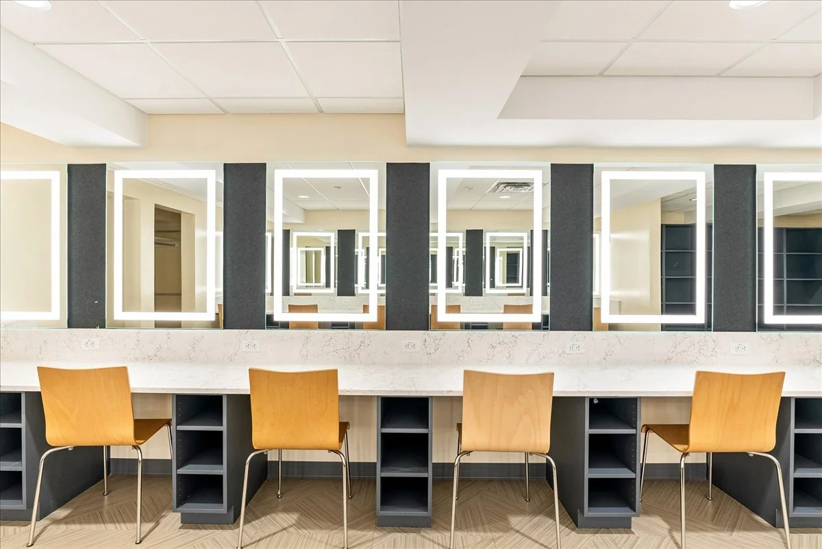 A modern salon or makeup station with four wooden chairs in front of a long white marble counter, illuminated rectangular mirrors with LED lighting, and dark shelving units underneath.