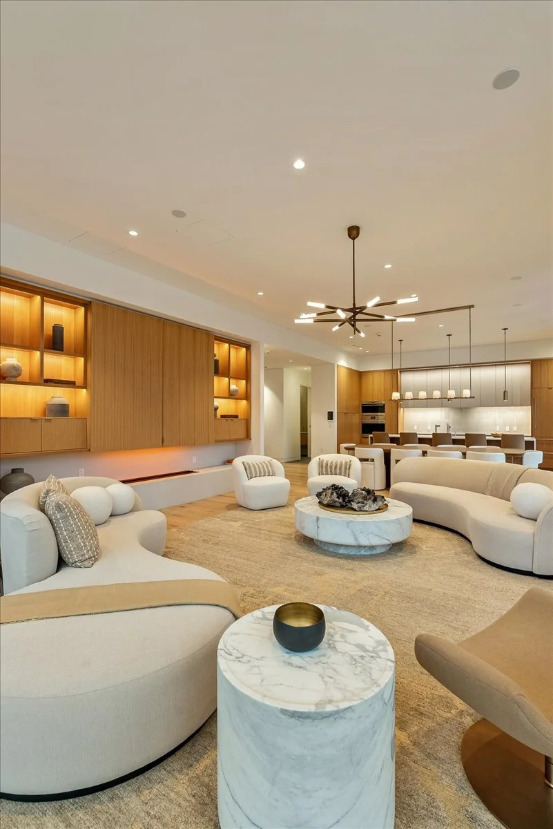 Modern living room with white curved sofas, armchairs, marble coffee tables, and built-in wooden wall shelves with decorative objects, open kitchen and dining area in background, and stylish lighting fixtures.