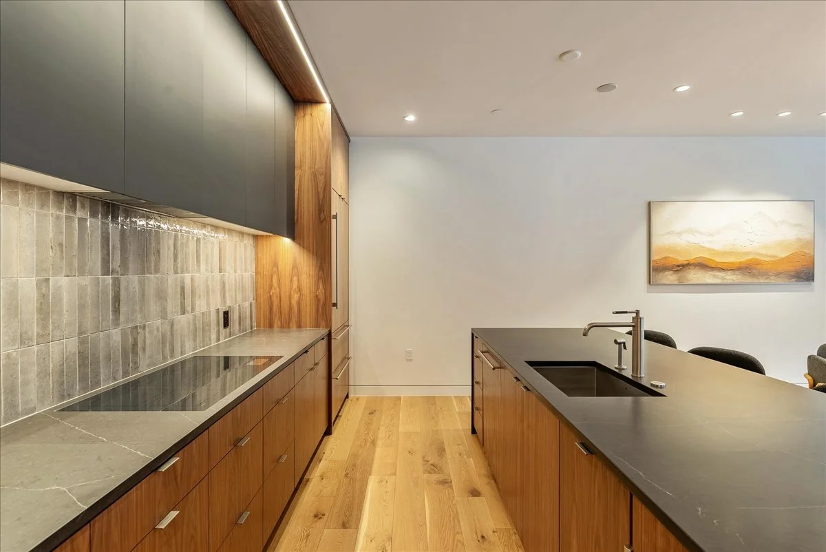 Modern kitchen with wooden cabinets, black countertop, gray tiled backsplash, and a white wall with a landscape painting.
