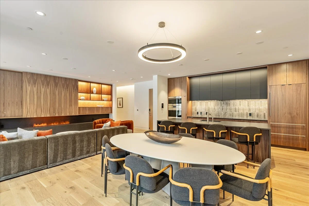 Modern open-plan kitchen and living room with wooden cabinetry, a large dining table with chairs, and a circular ceiling light fixture.