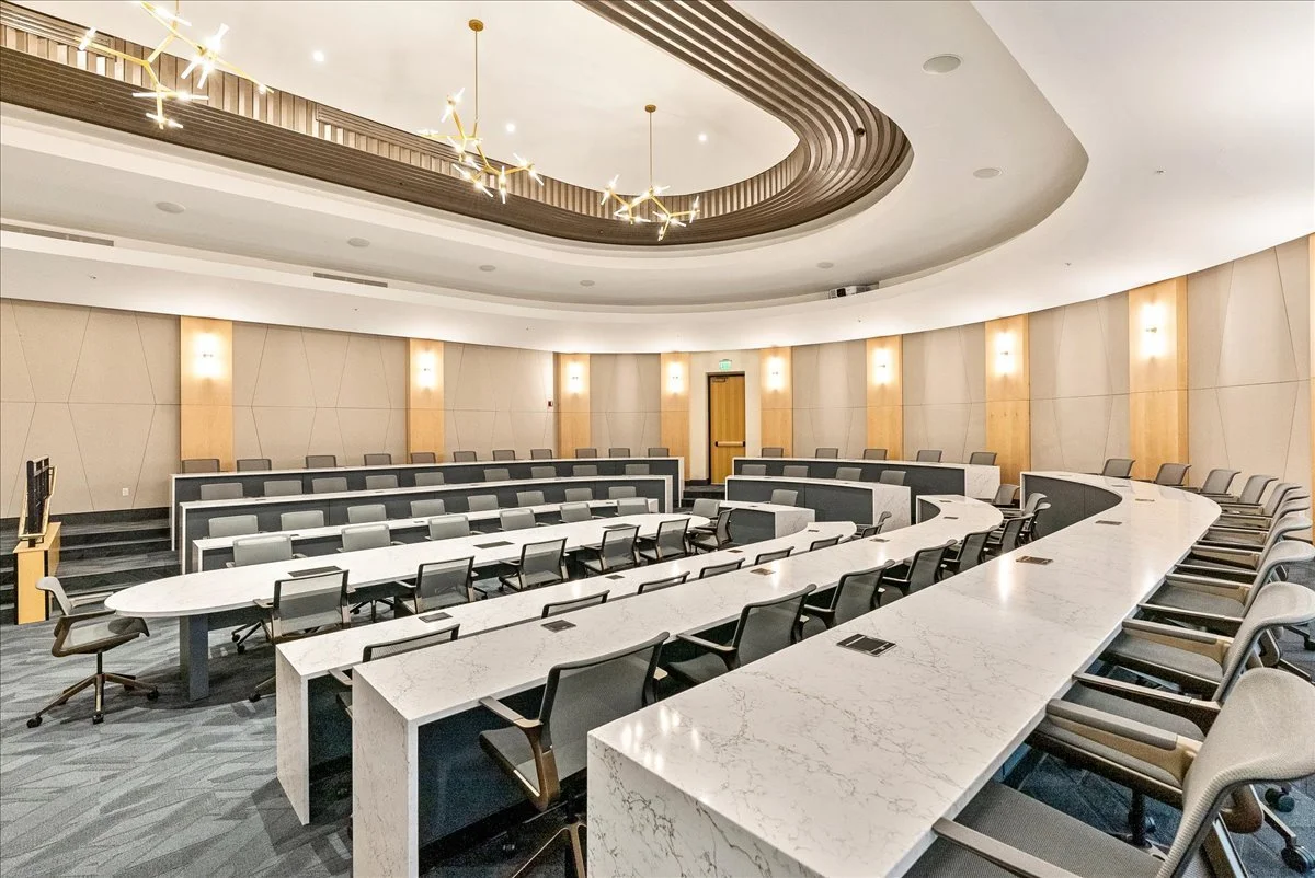 Empty conference room with curved white marble tables, ergonomic chairs, beige walls with vertical wooden accents, and a modern light fixture on the ceiling.