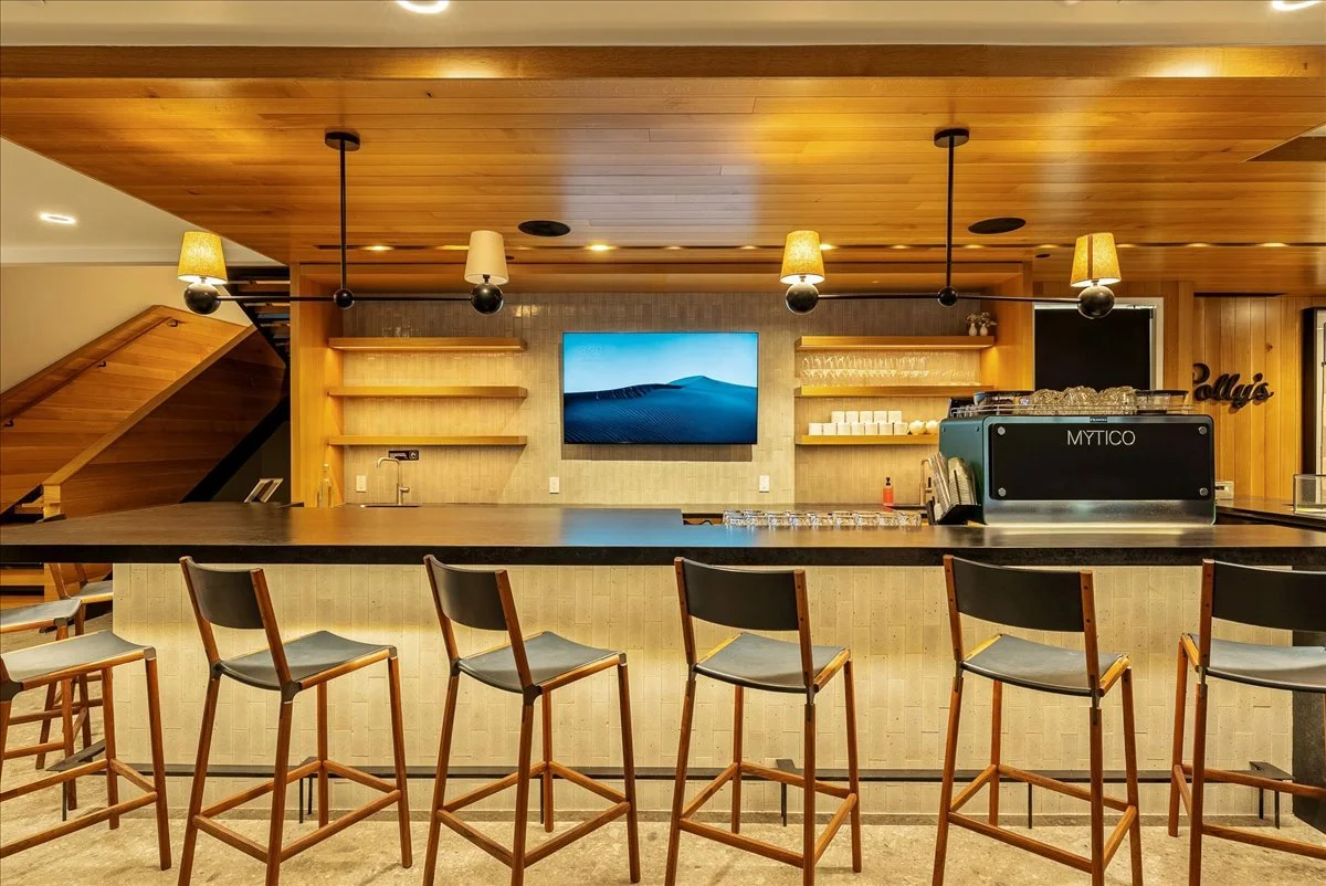 Empty bar with a row of five wooden chairs with black cushions, a black countertop, shelves behind, a TV in the center, and pendant lights hanging from a wood-paneled ceiling.