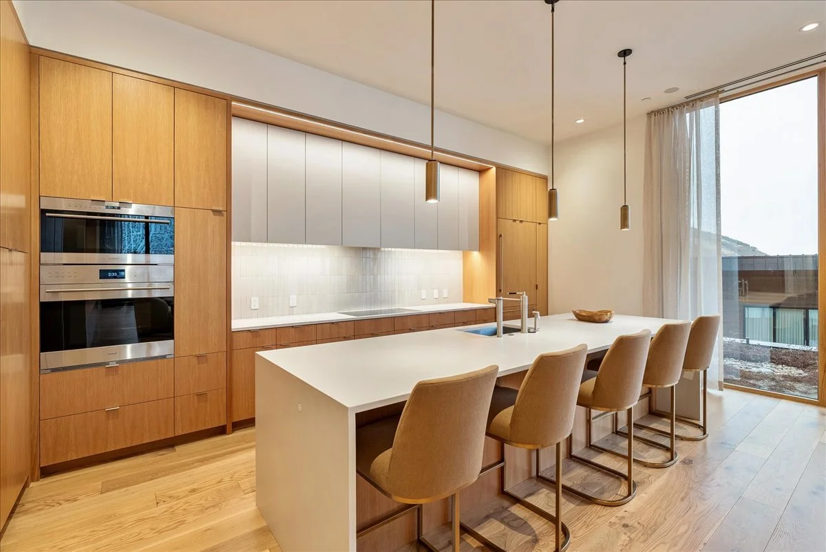 Modern kitchen with wooden cabinetry, white countertops, a large white island with beige chairs, stainless steel appliances, pendant lighting, and a large window with sheer curtains.