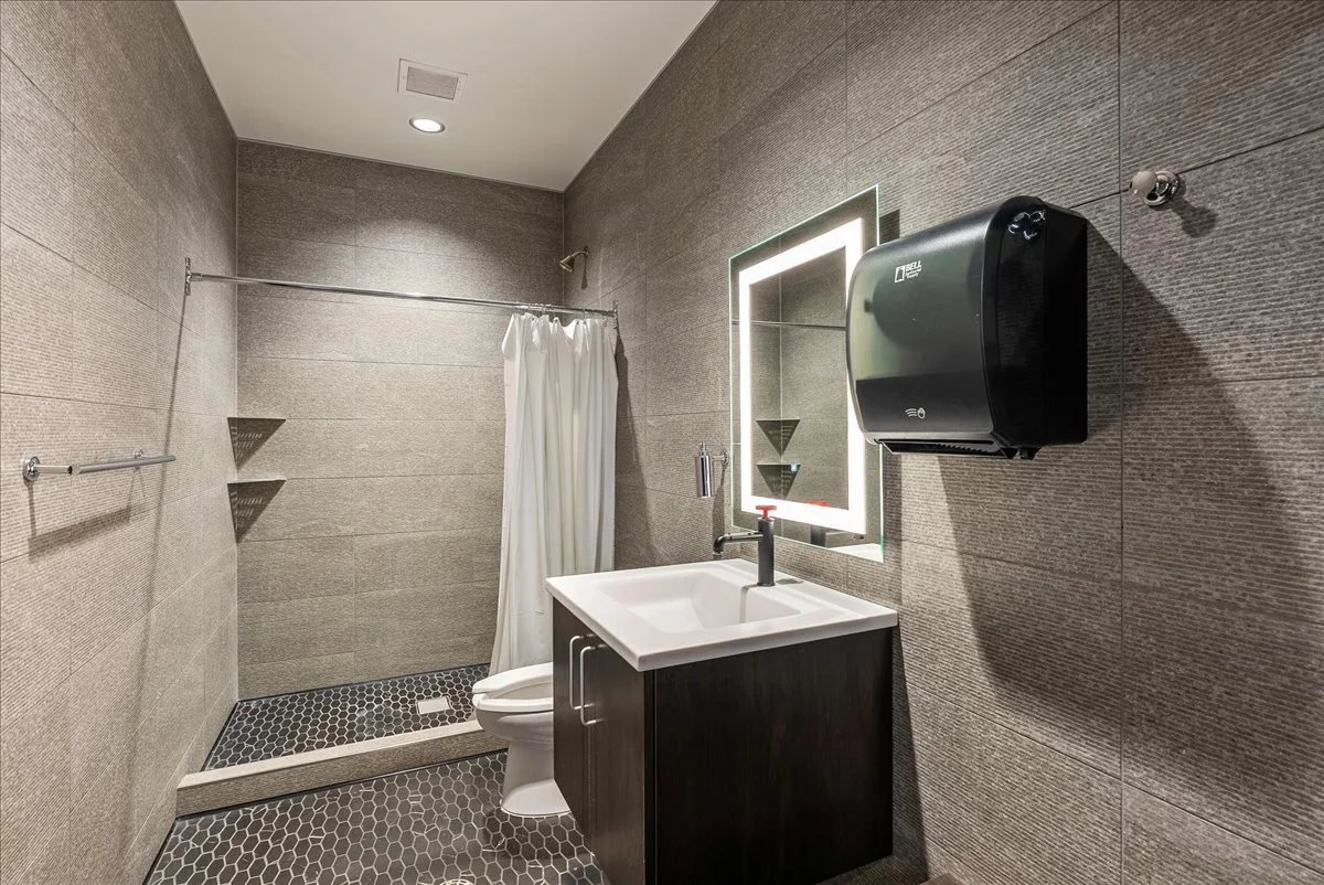 Interior of a modern bathroom with gray tiled walls and floor, a small white sink with a dark cabinet, a rectangular backlit mirror, a wall-mounted hand dryer, and a shower area with a curtain and corner shelves.