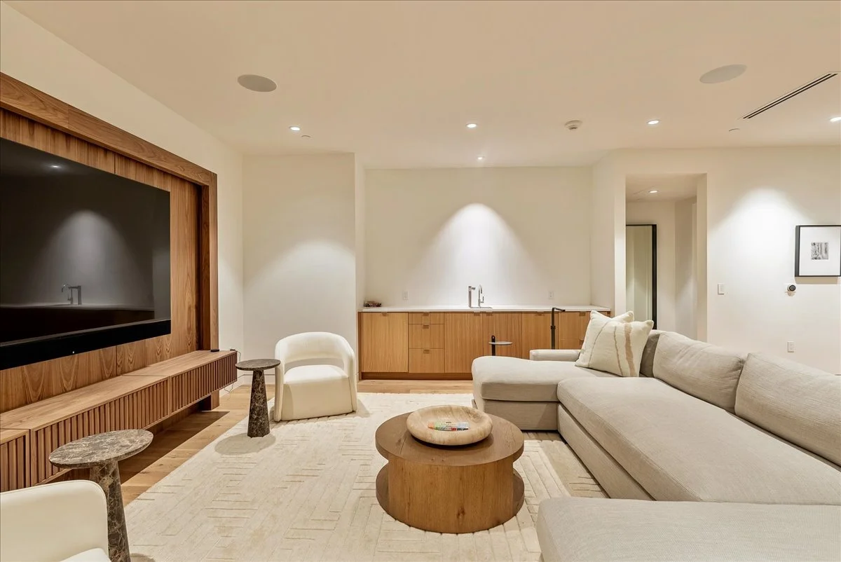 Modern living room with a large beige sectional sofa, two white armchairs, a wooden coffee table, and marble side tables, with a large flat-screen TV mounted on a wooden wall panel.