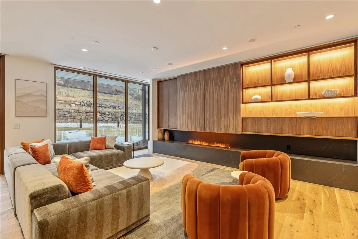 Modern living room with large windows, sectional sofa with orange and beige pillows, two burnt orange velvet armchairs, a round coffee table, a wall with wooden cabinets and built-in fireplace, decorative vases on wooden shelves, and a landscape view outside.