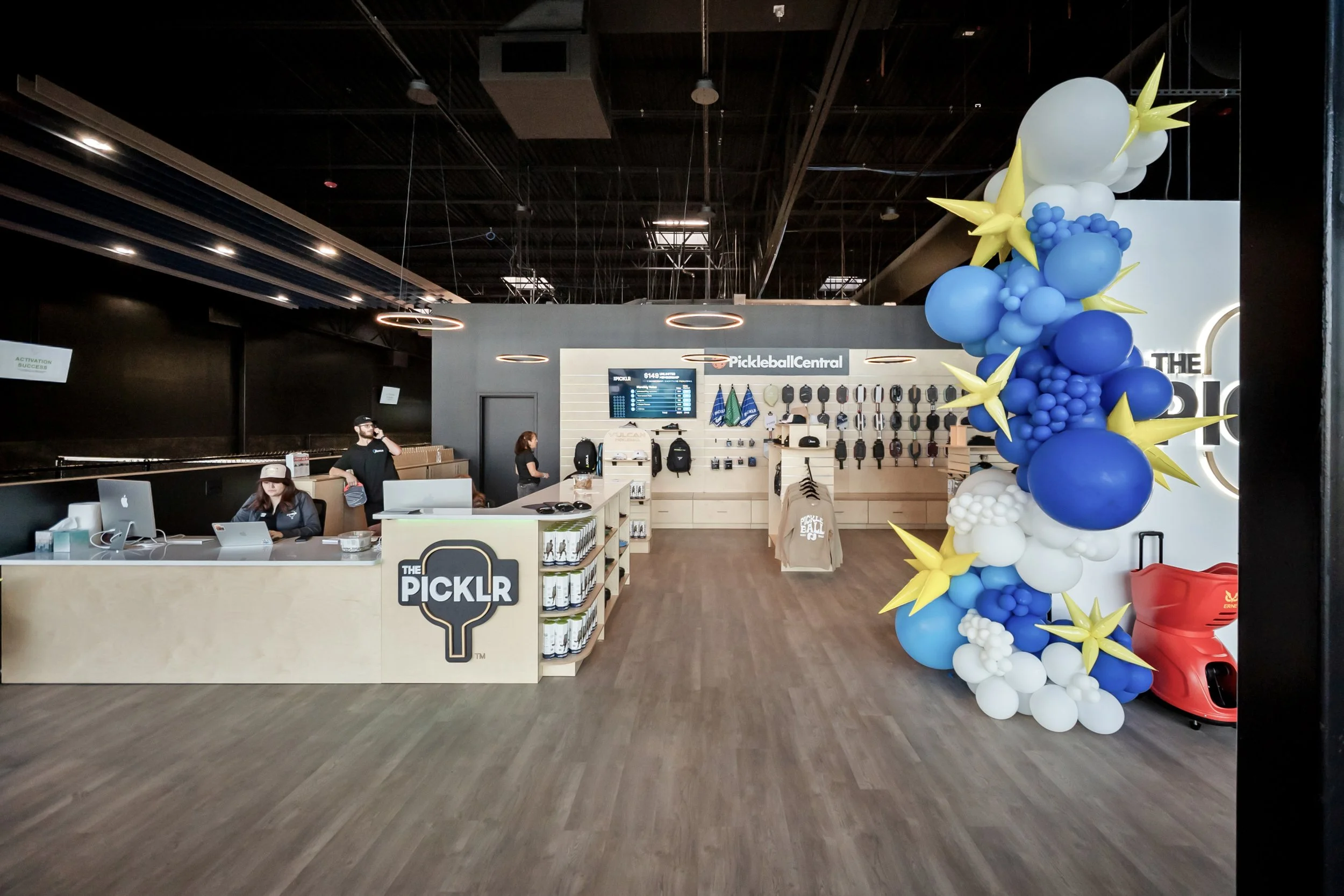 Interior of a store called The Picklr with a counter manned by staff, a colorful balloon decoration on the right side, and merchandise displayed on the wall, including backpacks and paddle rackets.