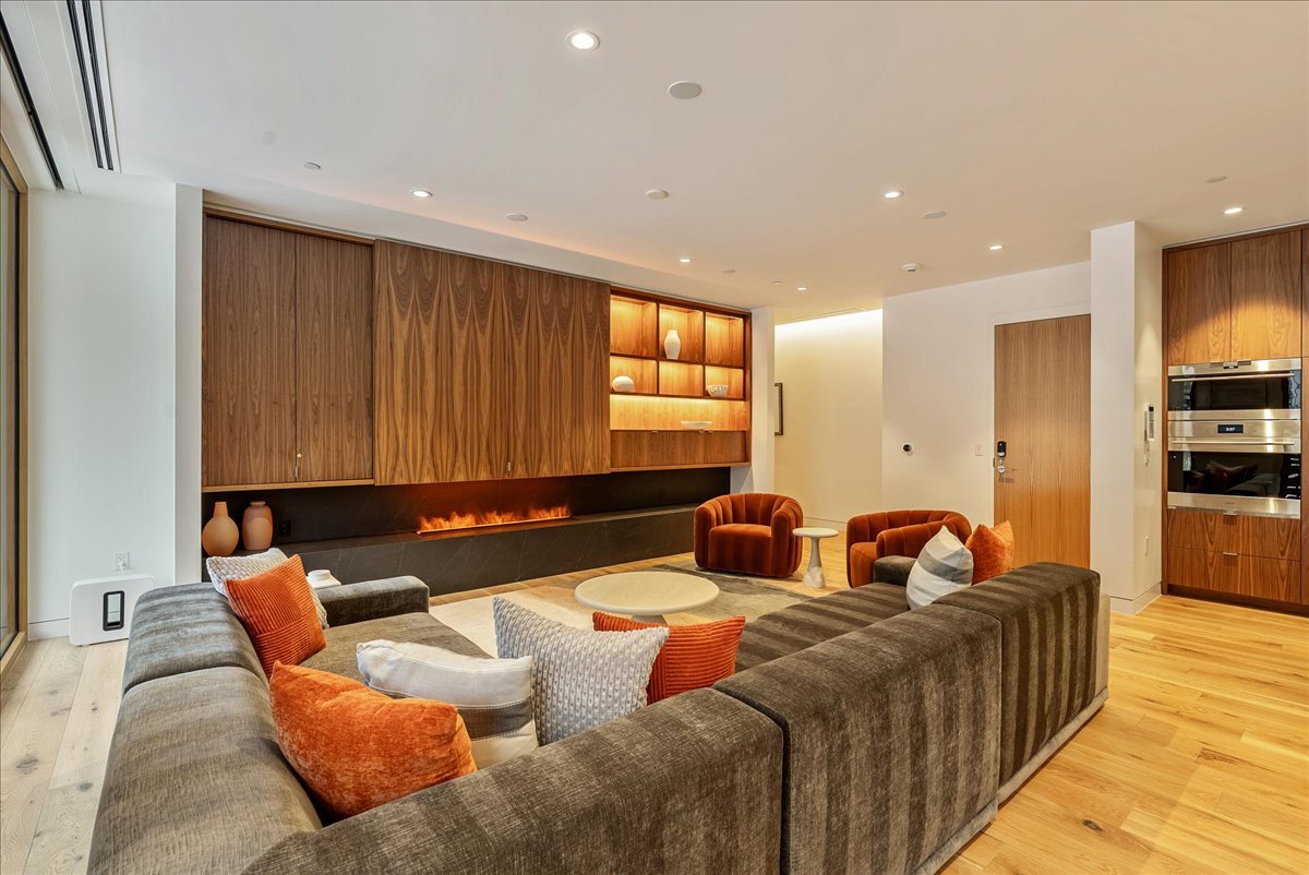 Modern living room with a large L-shaped gray sofa, orange and white throw pillows, and a circular coffee table. There are two rust-colored armchairs, a small side table, built-in wooden cabinet with decorative vases, and a linear fireplace. The room