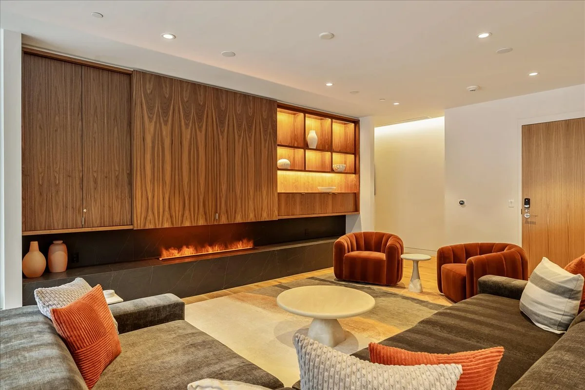 Living room with a sectional sofa, two red velvet armchairs, and a coffee table. Wooden cabinets and shelves with decorative items on the wall, and soft lighting.