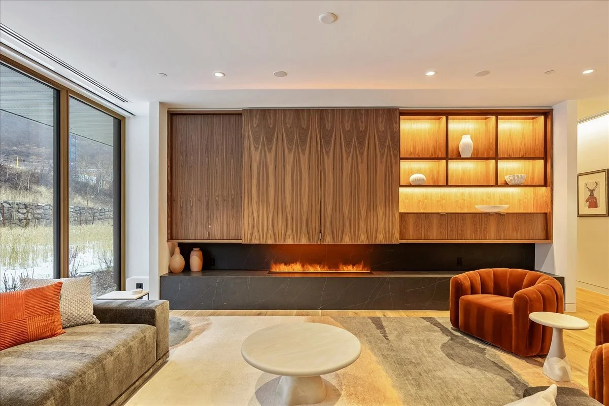 Modern living room with a large window, wooden wall with warm lighting, built-in fireplace, and contemporary furniture including a gray sofa with red pillows, orange velvet armchairs, a round coffee table, and open shelving with decorative objects.