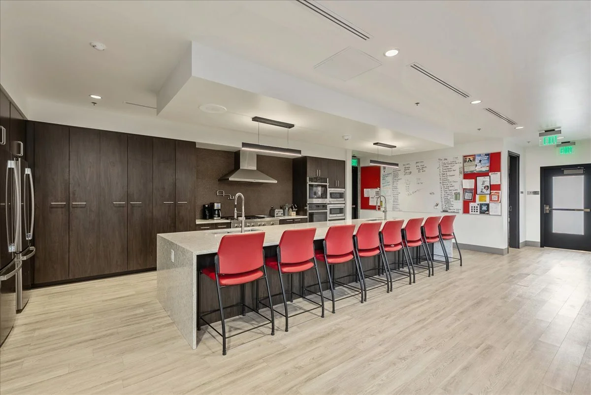 Modern kitchen area with a long counter and nine red chairs, equipped with appliances, cabinets, and a whiteboard on the wall.