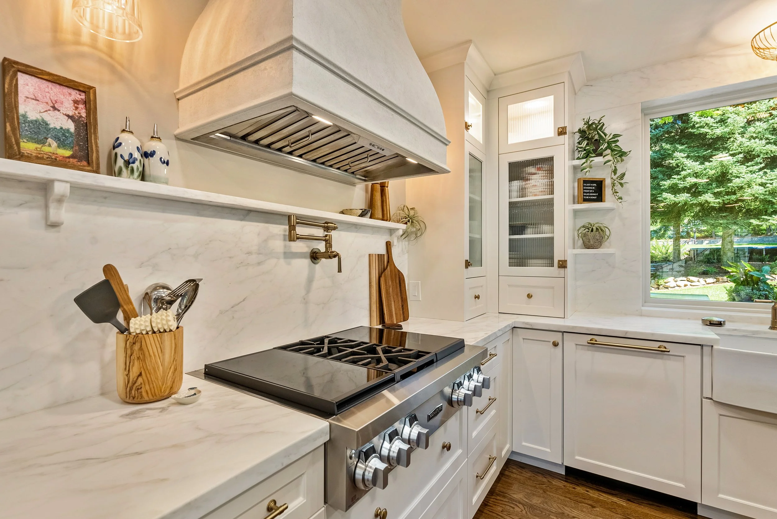 A modern kitchen with white marble countertops and backsplash, stainless steel gas stove, wooden utensils in a holder, framed picture, decorative vases, plant, and large window showing green outdoor scenery.