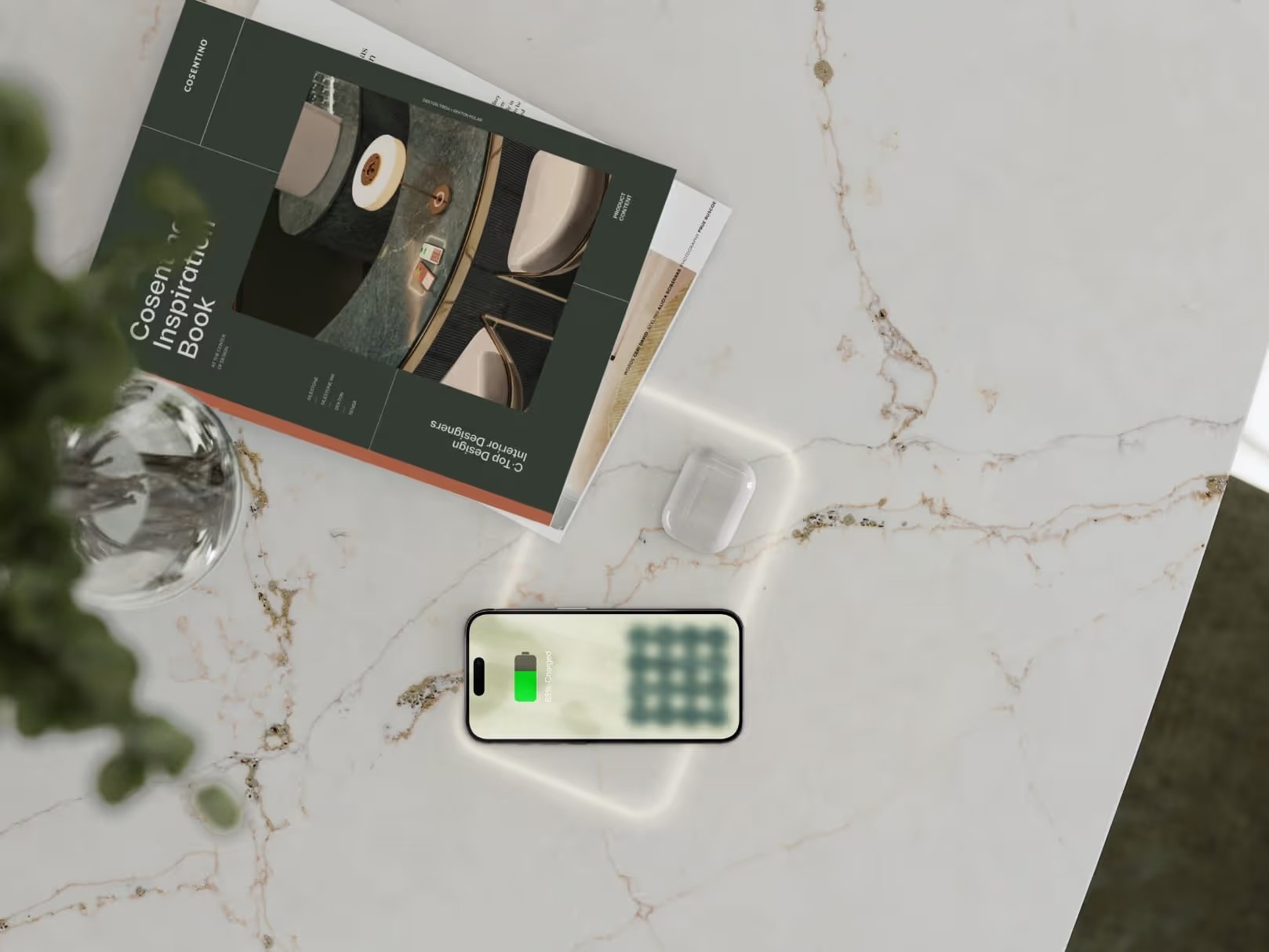 A marble countertop with a magazine titled "Cosentido Inspiration Book," a glass with water, a pair of wireless earbuds in a case, and a smartphone displaying a full green battery charge. Part of a plant is visible on the left.