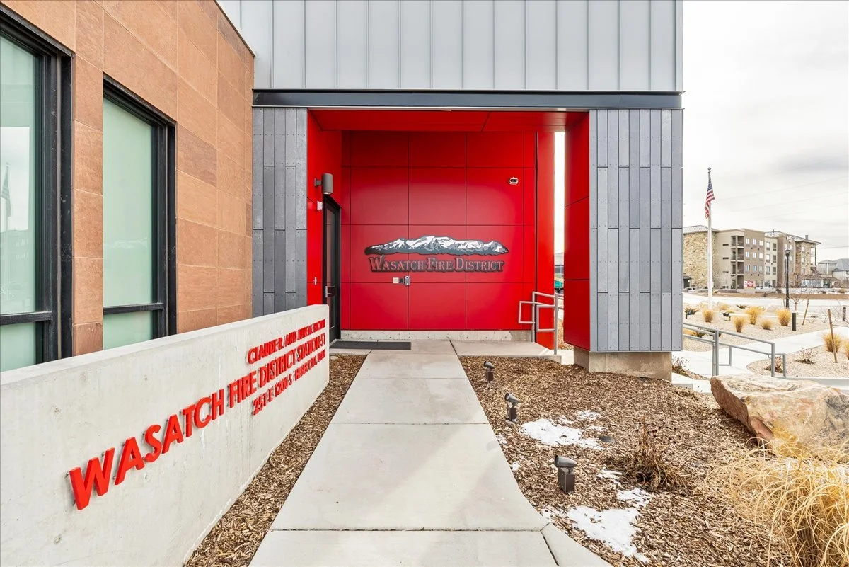 Entrance to the Wasatch Fire District building with a red wall featuring a mountain logo and sign, and a sidewalk leading to the door. There is a concrete wall with red text on the left, and a small landscaped area with rocks and sparse plants on the right.