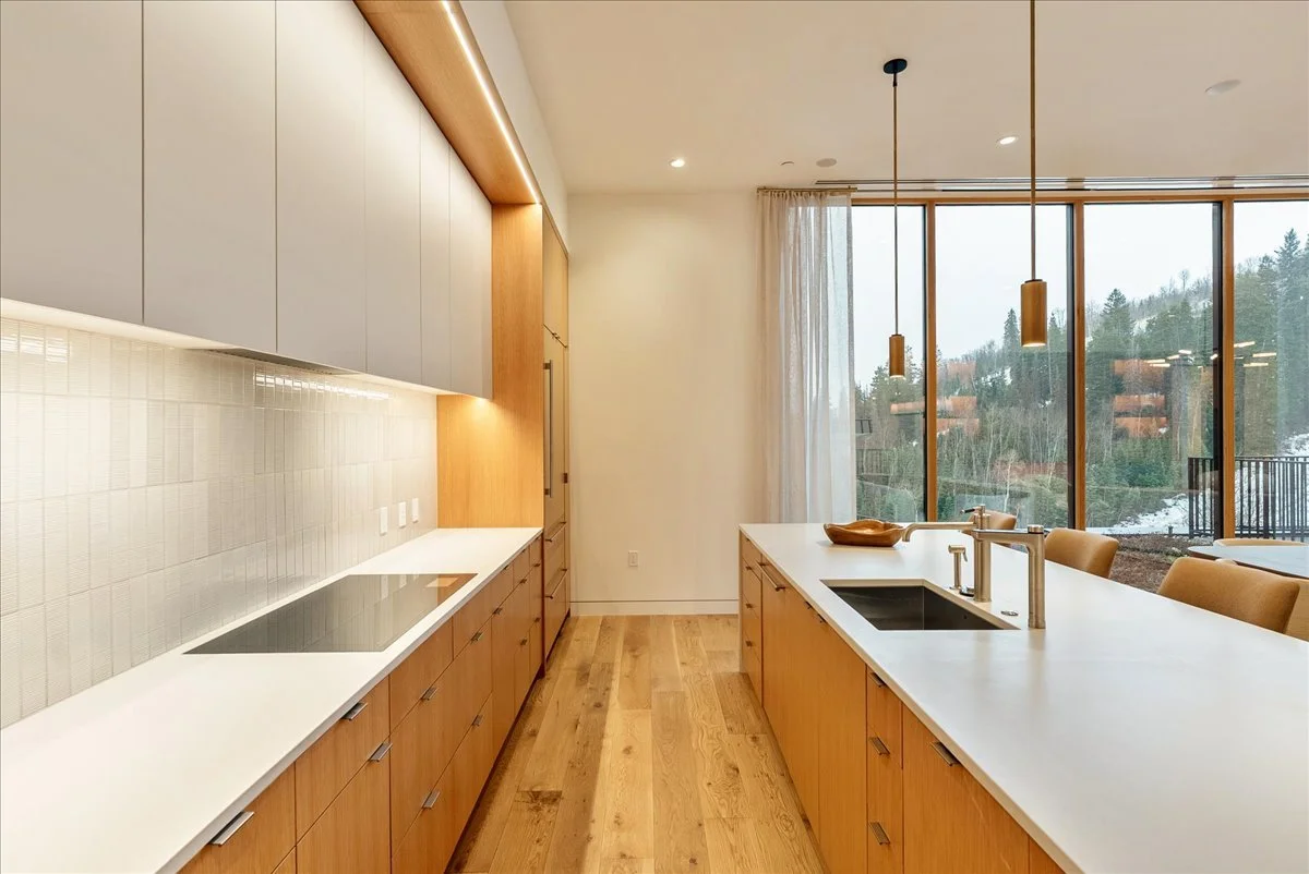 Modern kitchen with wooden cabinetry, white countertops, an electric stovetop, a black sink, and a large window with curtains overlooking a wooded landscape.