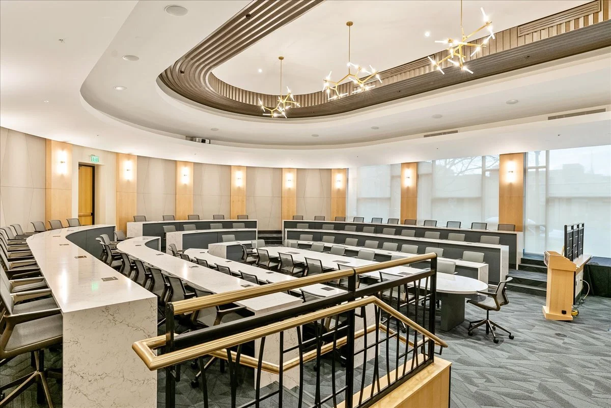 Empty modern conference room with tiered seating, desks, and chairs, illuminated by ceiling lights and decorative hanging fixtures.