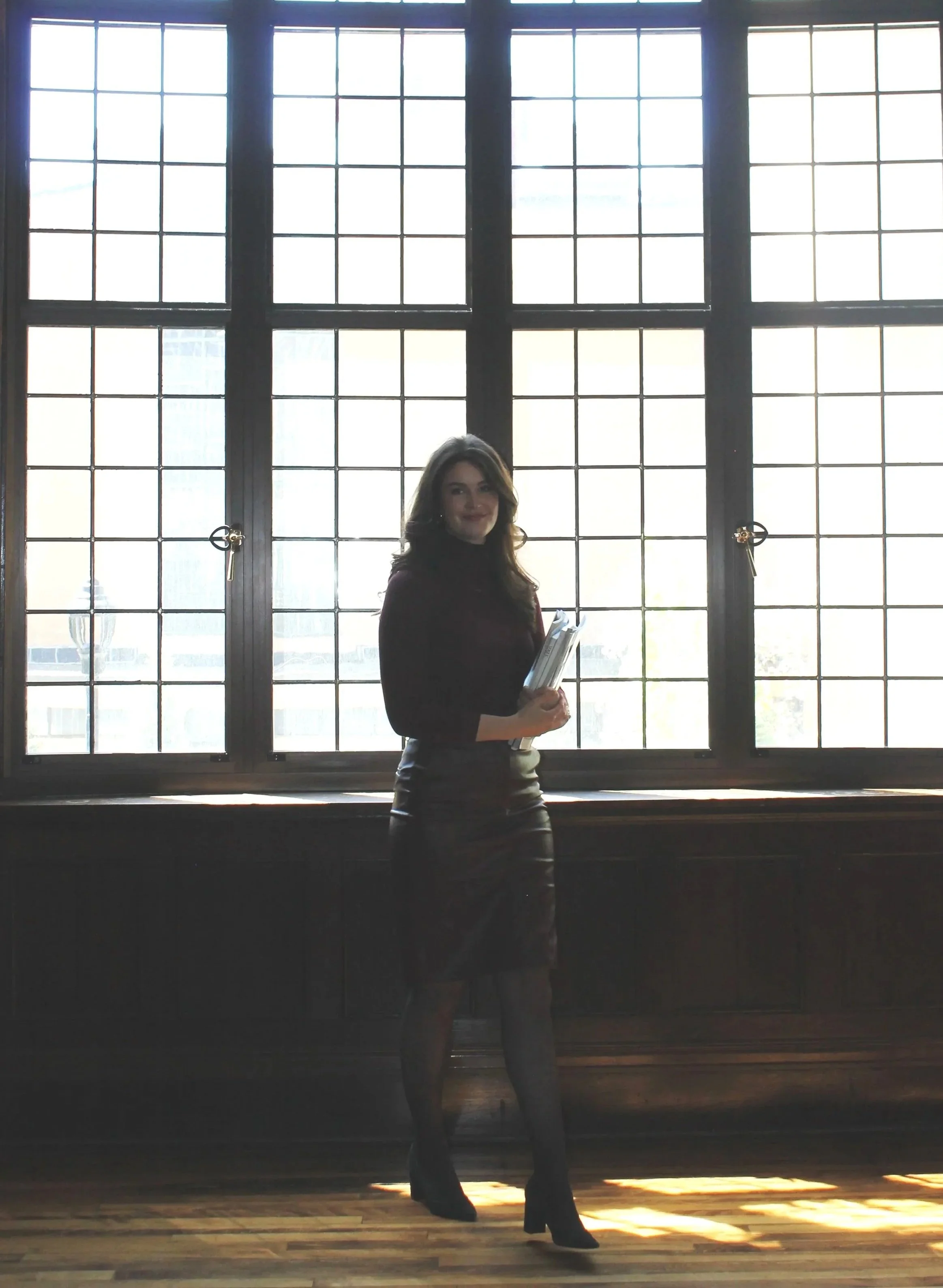 A woman standing indoors in front of large window with grid pattern, holding papers, wearing dark clothing and high-heeled boots, with sunlight casting shadows on the wooden floor.