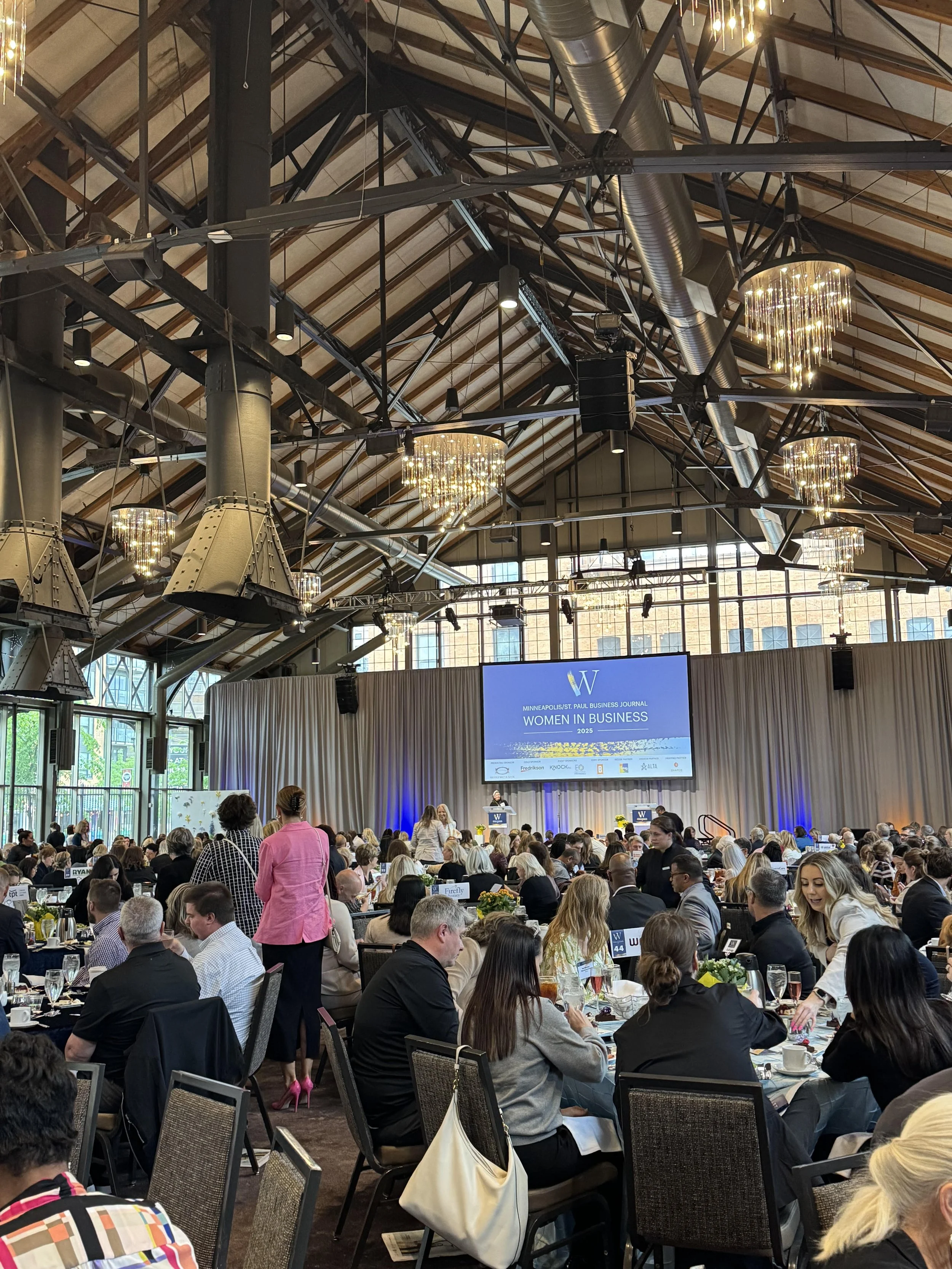 A large conference or banquet hall filled with many attendees seated at round tables. There is a stage at the front with a large screen displaying 'Women in Business 2025,' and multiple chandeliers hang from the high industrial-style ceiling with exposed ductwork and wooden beams. People are engaged in conversation and dining.
