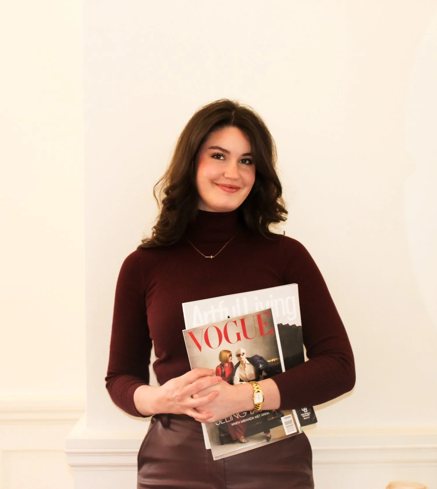 A woman with shoulder-length brown hair smiling, holding a Vogue magazine while standing against a plain beige wall.