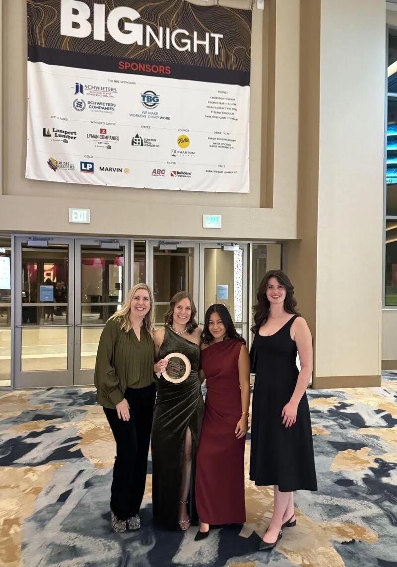 Four women dressed in formal attire standing together in front of a large event banner, smiling for a photo at an indoor event.