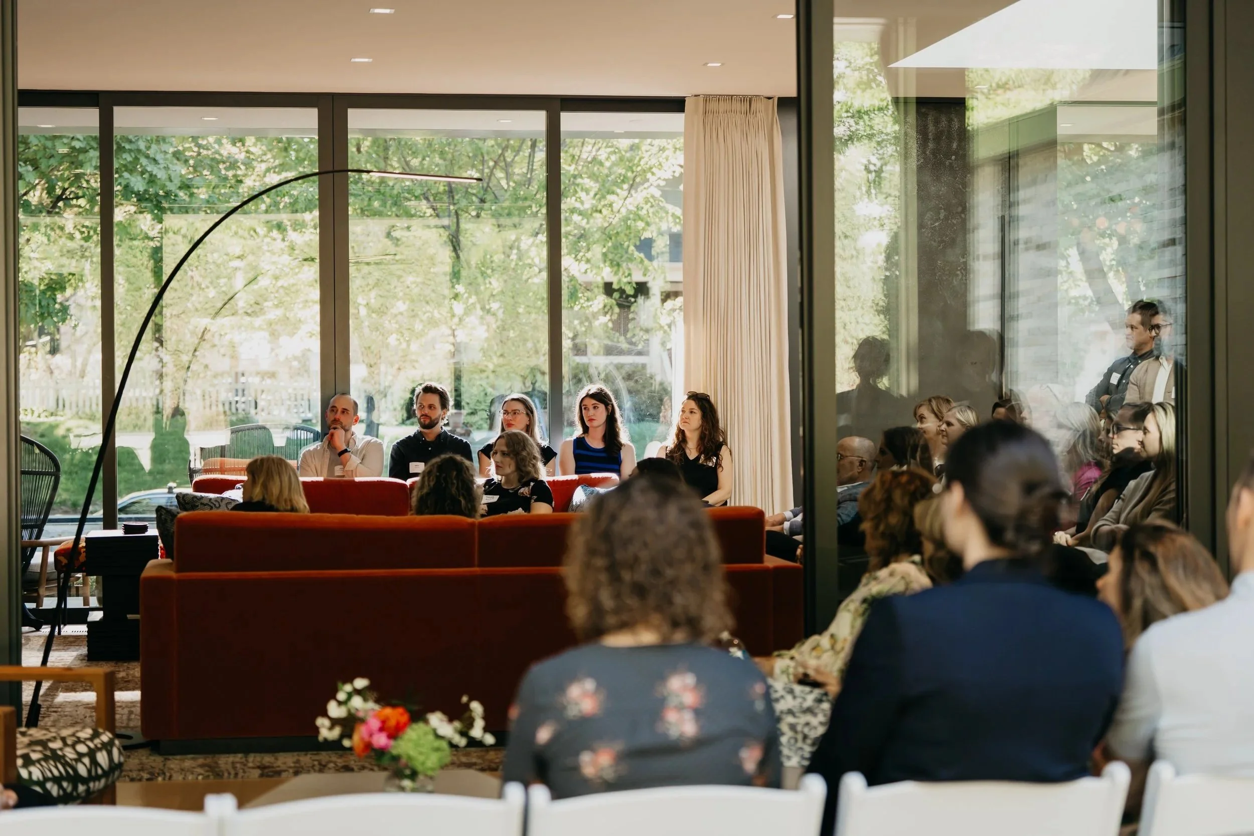 People seated in a room during a presentation or event, with large windows showing a green outdoor scene.