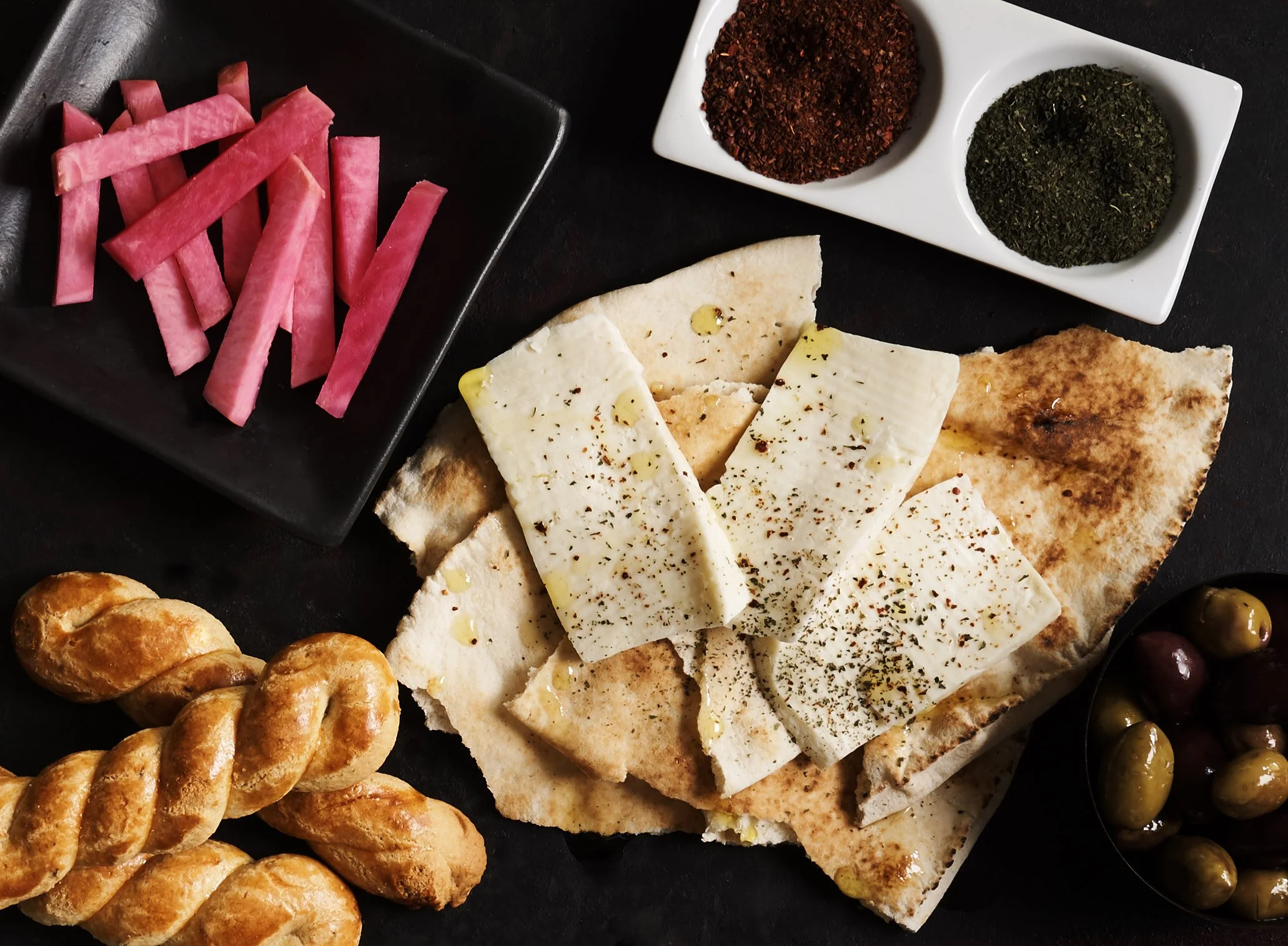 A tray of assorted foods including sliced watermelon radish, various spices in white dishes, wedges of cheese, flatbread, and small bread rolls.
