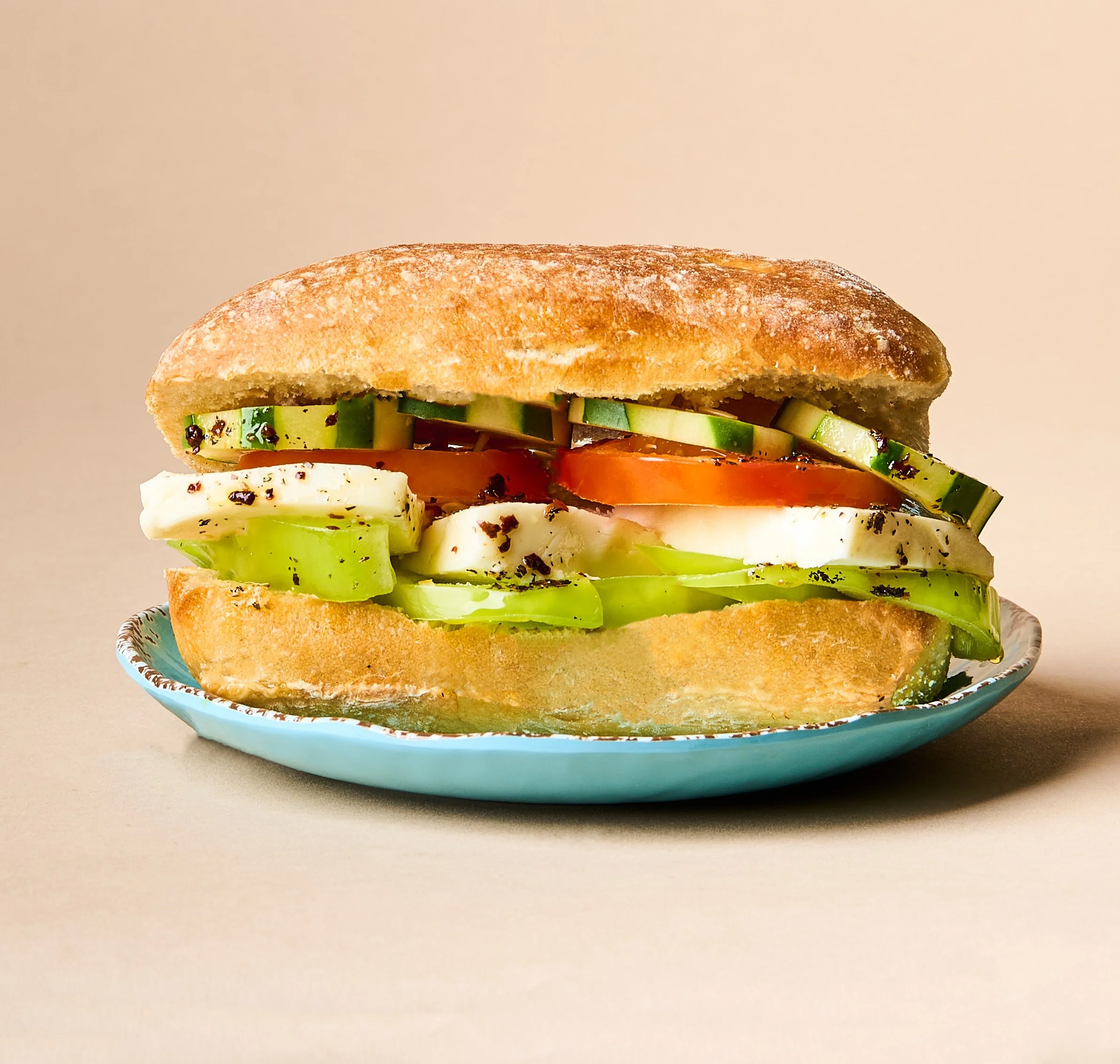 Close-up of a sandwich with slices of cucumber, tomato, and cheese on a toasted bun, garnished with black pepper, on a decorative plate.