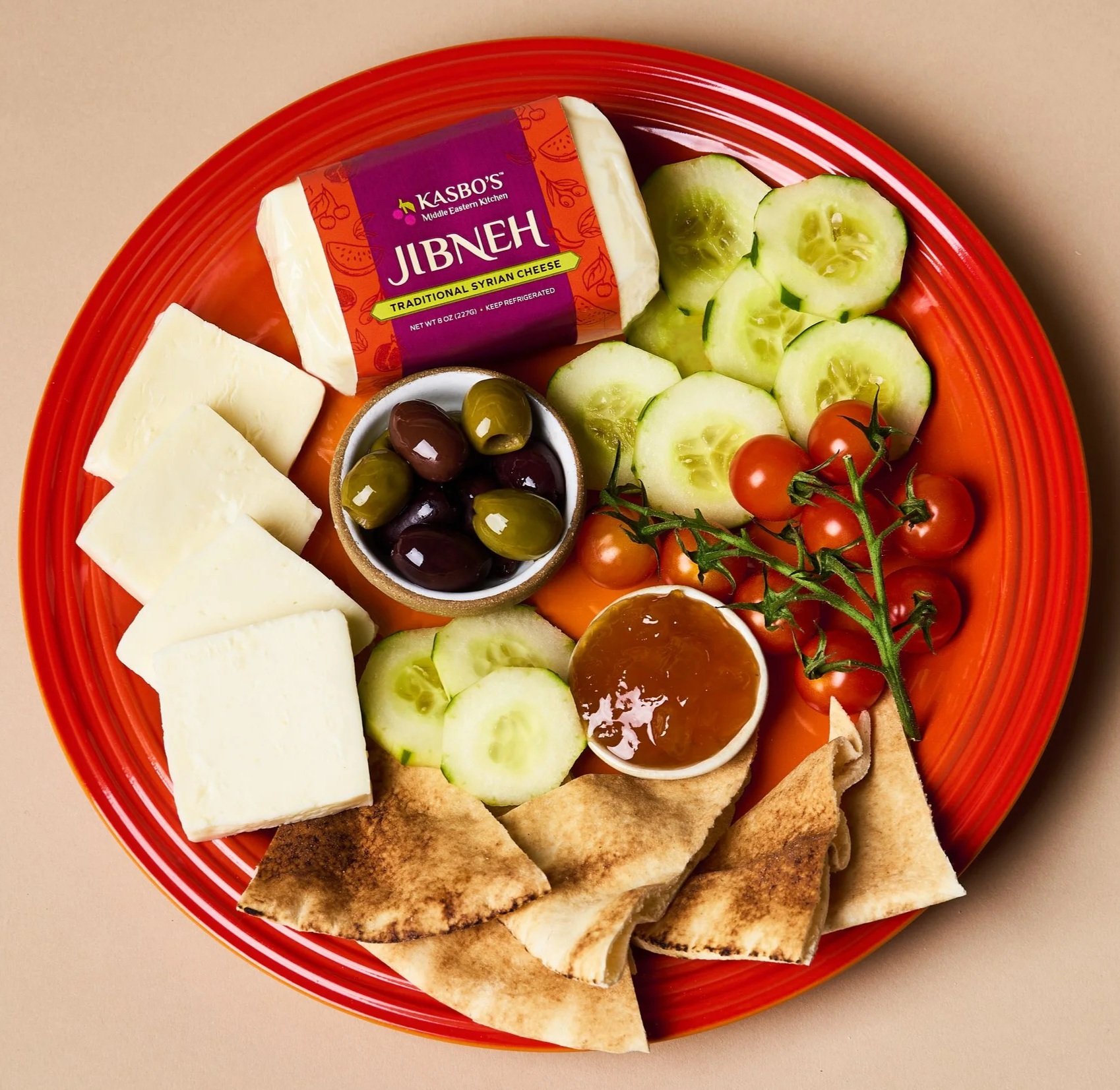 A red plate with slices of cheese, cucumber, cherry tomatoes on the vine, green and purple olives in a small bowl, pita bread, and a block of Syrian cheese labeled 'Jibneh' from Kasbo's Middle Eastern Kitchen.