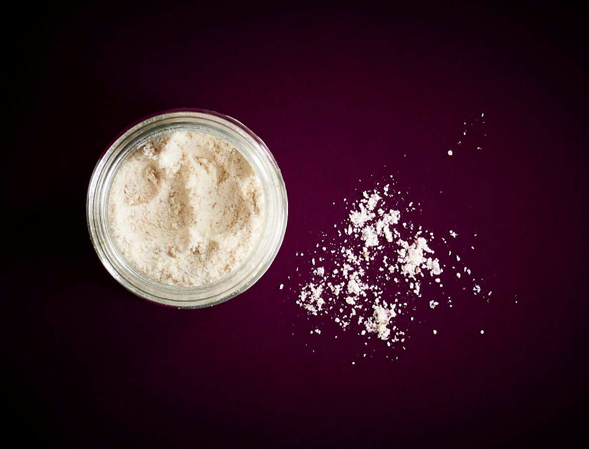 A glass white powdered cherry pits called mahleb with some sprinkled onto a dark purple surface.