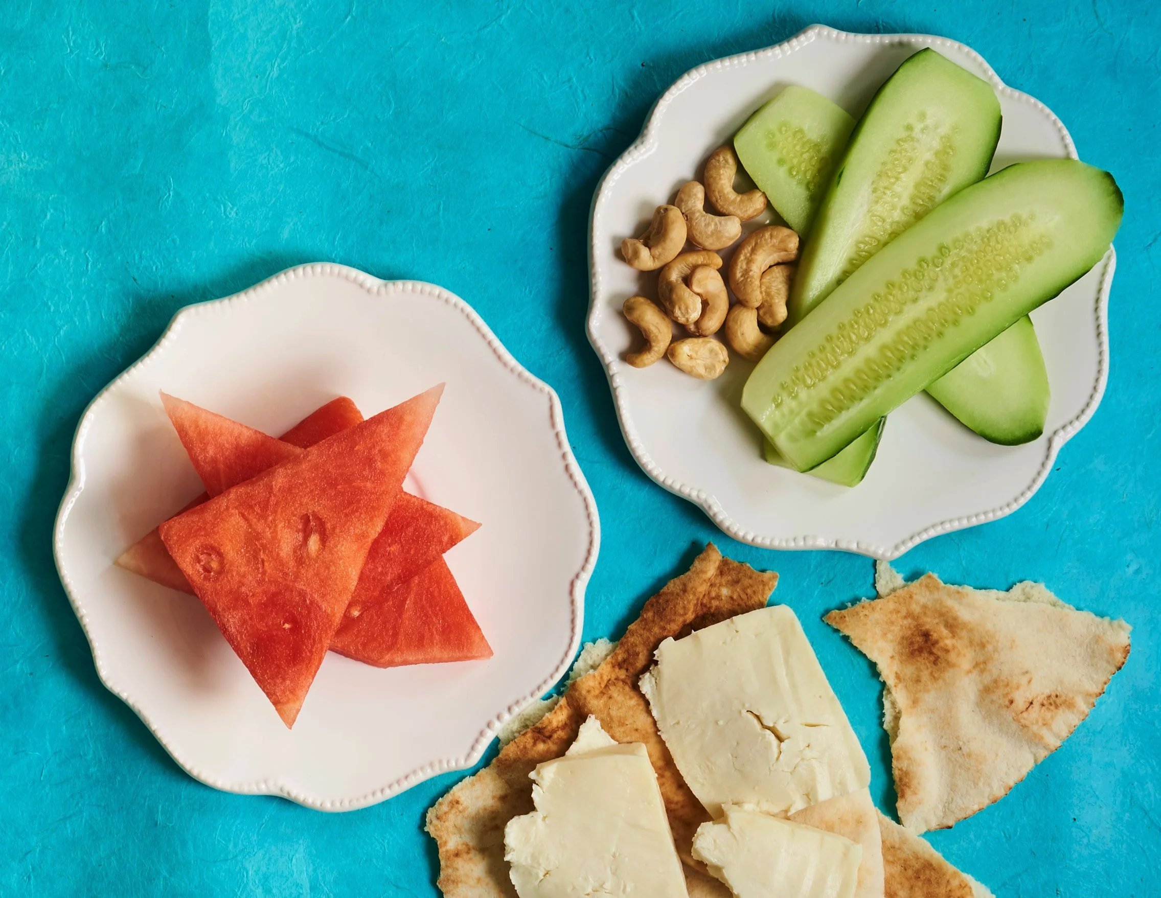A white decorative plate with sliced cucumbers, watermelon, paired with jibneh, syrian cheese, and cashews, on a blue surface.