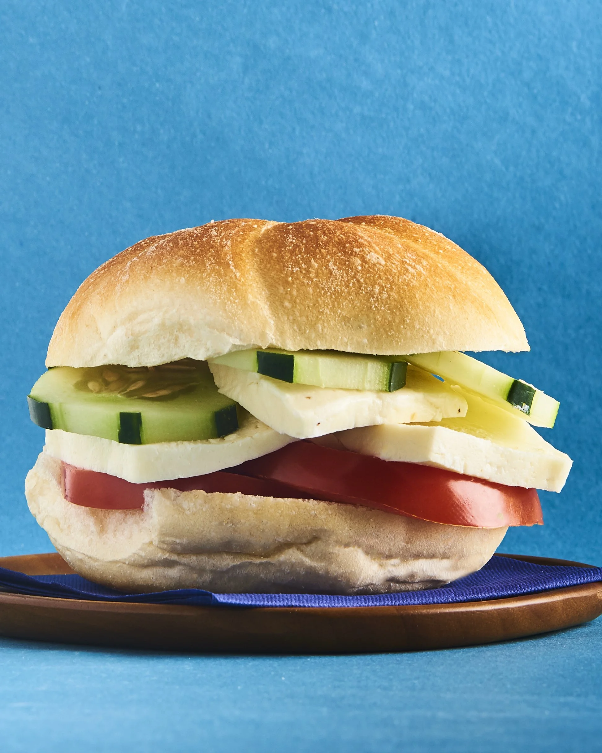 Close-up of a sandwich with layers of cucumber, tomato, and jibneh, syrian cheese on a bun, placed on a blue plate against a blue background.